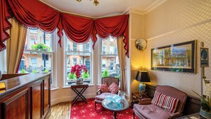 Habitación de esquina con escritorio de madera, alfombra roja con estampado, sofá y sillón con vistas a la ciudad a través de amplias ventanas.