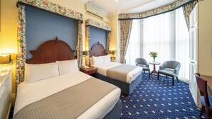 Una habitación de hotel con dos camas, una pequeña mesa con sillas y un ventanal con cortinas.