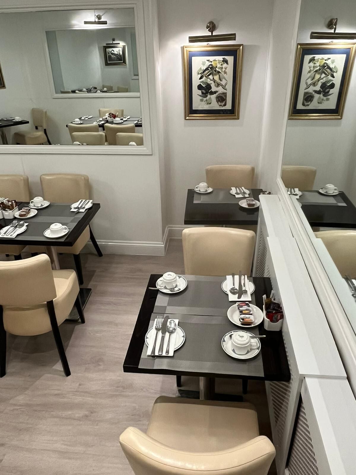 Un coin repas avec des tables dressées pour quatre personnes, chacune agrémentée d’une assiette blanche, de couverts et d’un petit dessert. La pièce présente un design moderne, avec des chaises de couleur beige et un grand miroir au mur.
