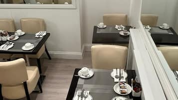 Un coin repas avec des tables dressées pour quatre personnes, chacune agrémentée d’une assiette blanche, de couverts et d’un petit dessert. La pièce présente un design moderne, avec des chaises de couleur beige et un grand miroir au mur.