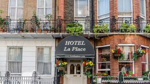 L’entrée d’un hôtel, avec une enseigne noire sur laquelle on peut lire « HÔTEL La Place » et l’adresse de son site web. Des jardinières remplies de fleurs sont posées sur les rebords des fenêtres, et une porte blanche, surmontée du numéro « 17 », donne accès à l’intérieur.