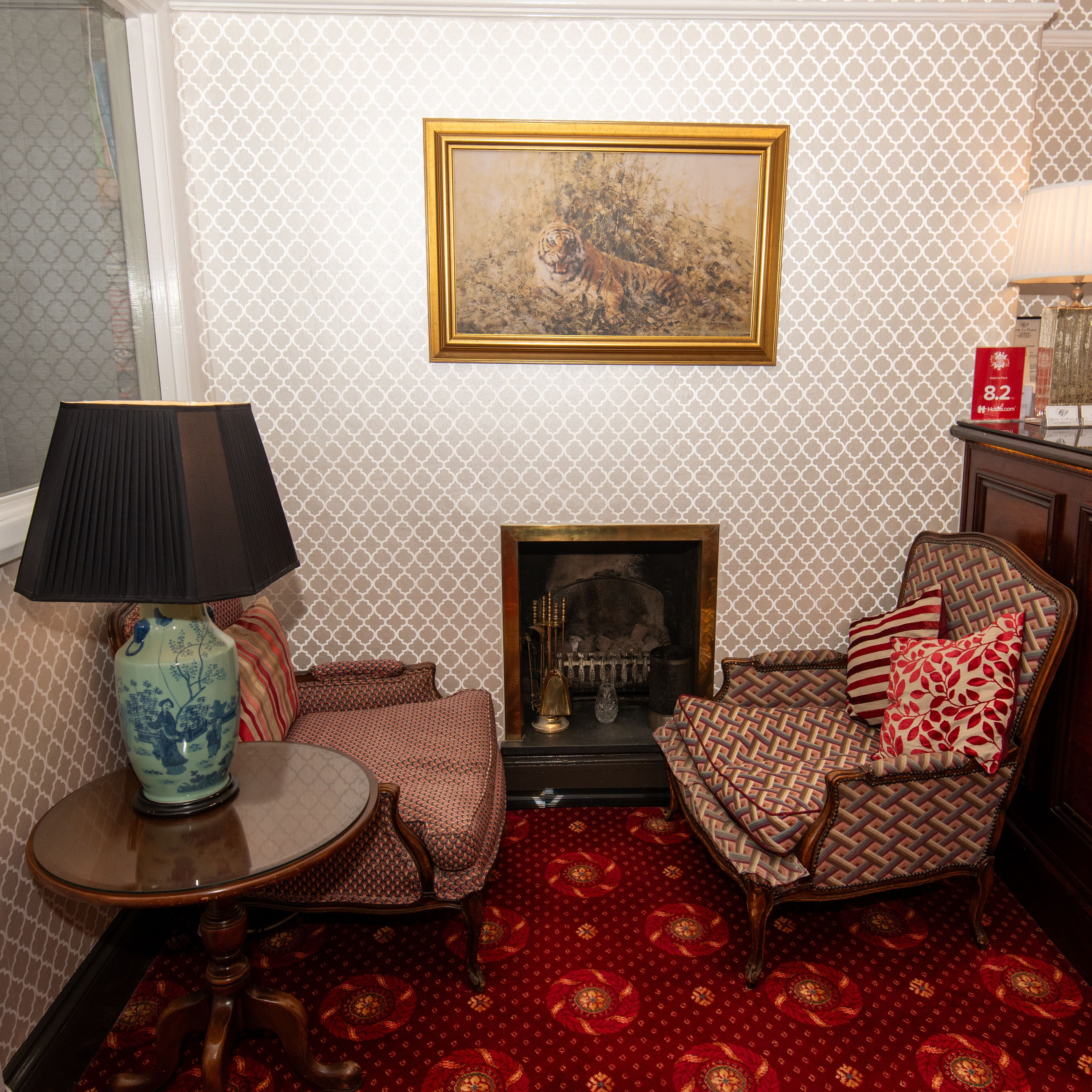 A room with patterned wallpaper, a framed painting, a fireplace, a sofa, a chair, a table with a lamp, and a decorative vase.