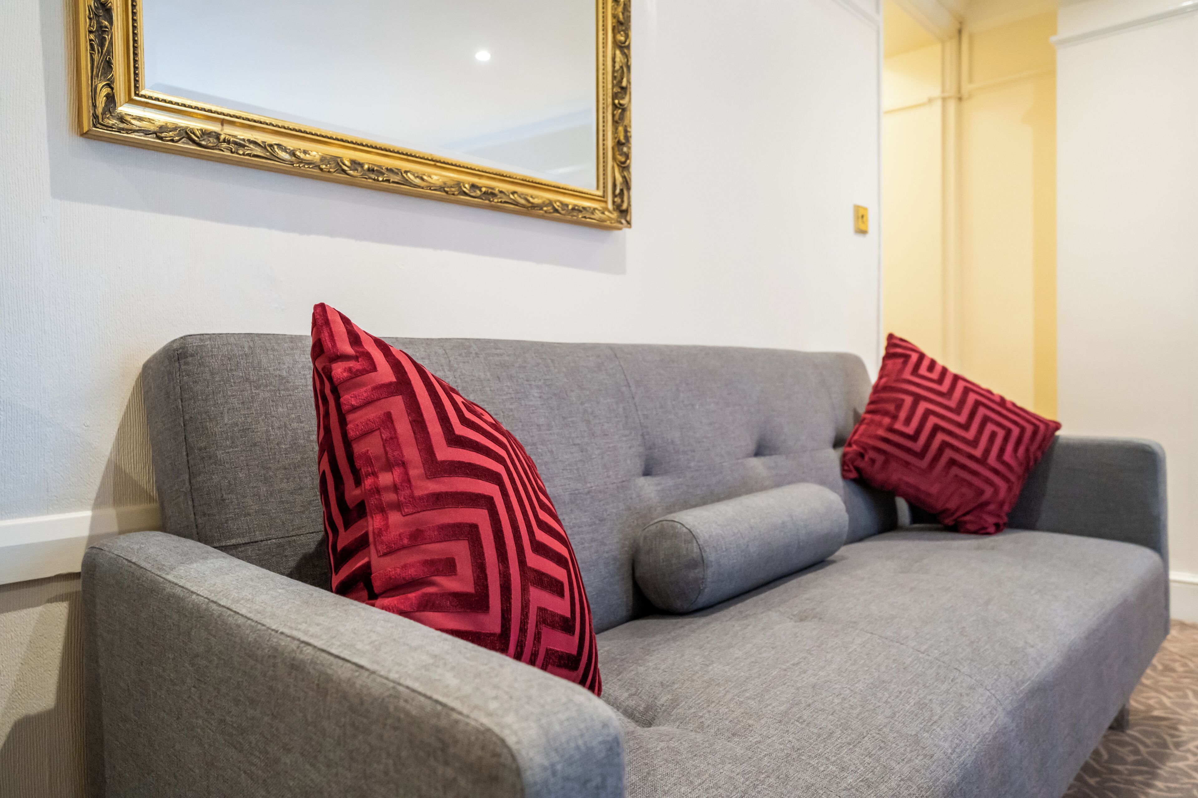 A grey sofa with two red patterned pillows and a gold-framed mirror on the wall.
