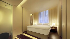 Habitación de hotel con una cama grande, una mesita de noche con lámpara, una ventana con cortinas traslúcidas y suelo de madera.