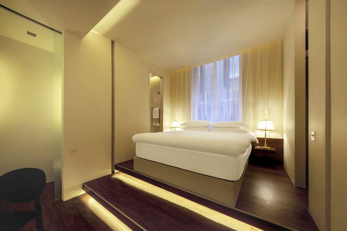 Habitación de hotel con una cama grande, una mesita de noche con lámpara, una ventana con cortinas traslúcidas y suelo de madera.