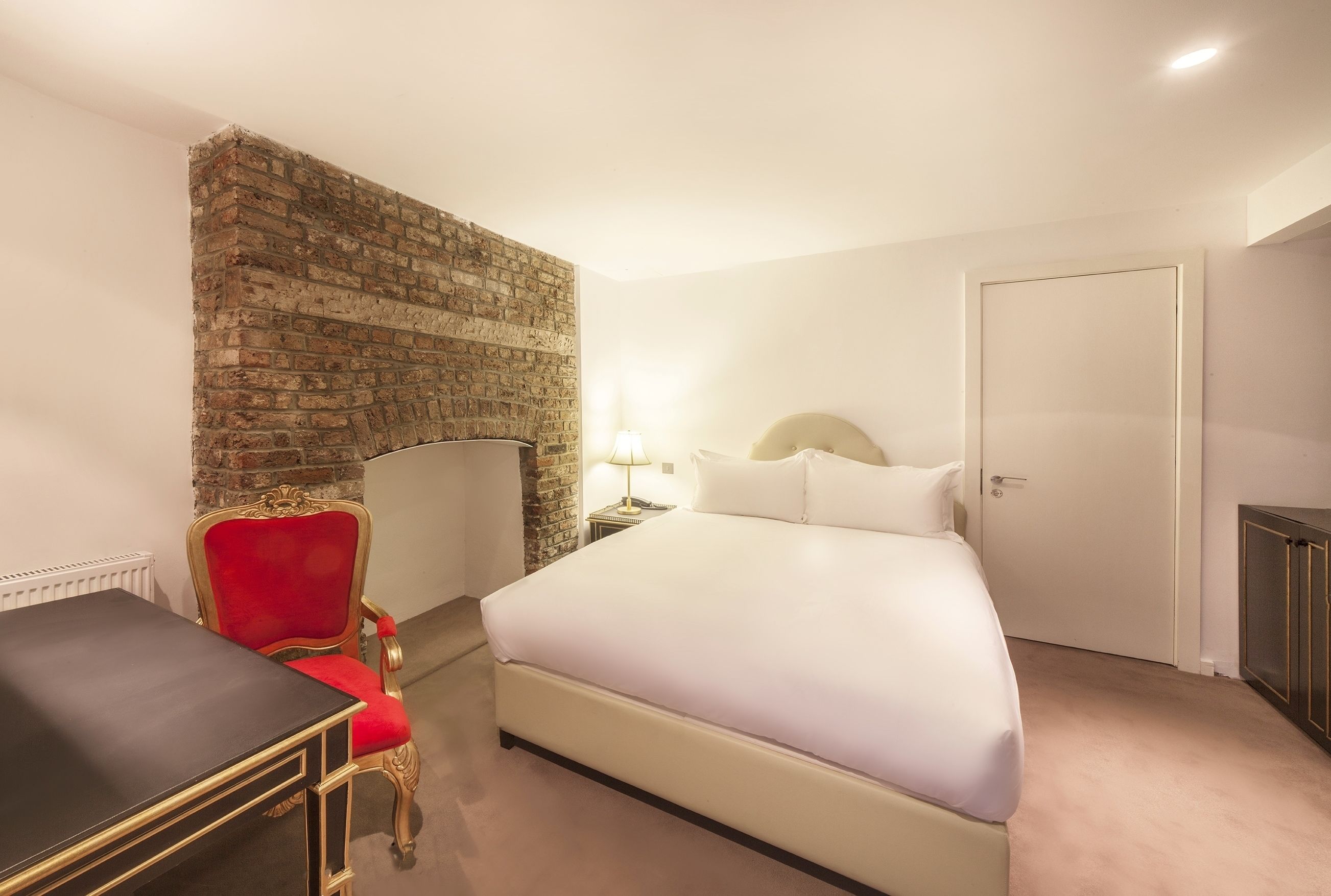 A hotel room with a brick accent wall, a bed with white linens, a red upholstered chair, a small table, and a nightstand with a lamp.