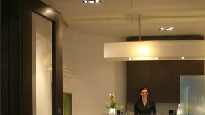 Zona de recepción con recepcionista, un mostrador, un área de asientos con un banco y una planta en maceta.