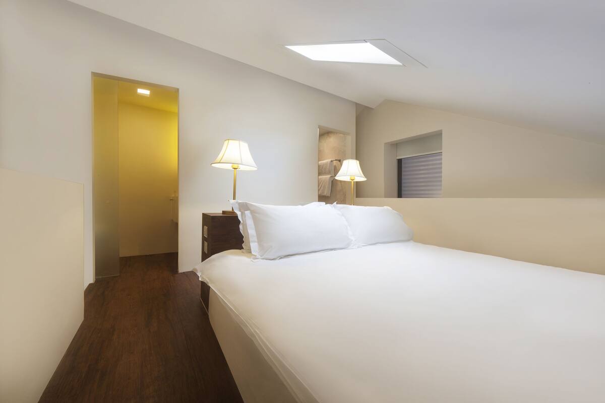 Habitación de hotel con cama, dos lámparas de noche, ventana con persianas, puerta que conduce a otra habitación y una lámpara en el techo.