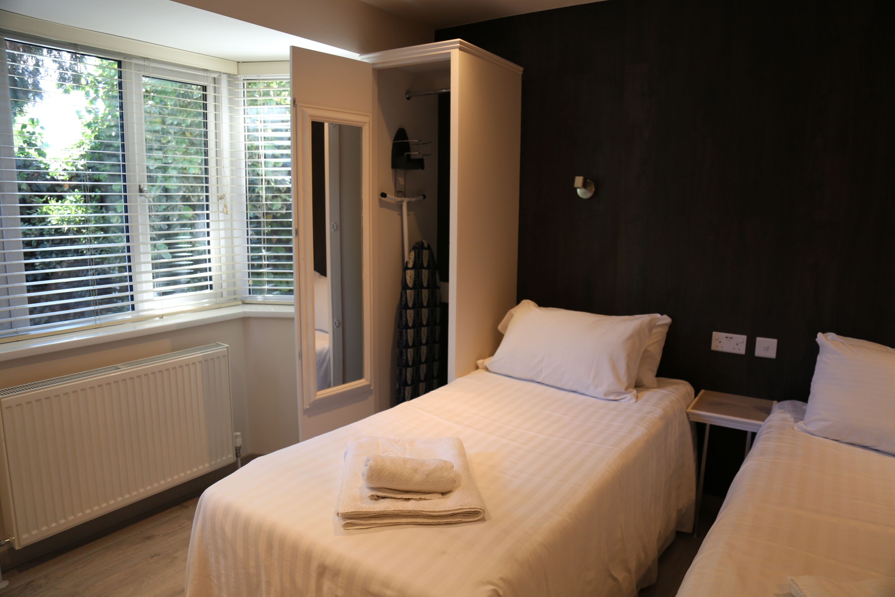 A hotel room with two beds, a window with blinds, a radiator, and a shower area.