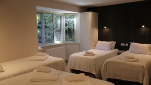 A hotel room with three single beds, each with a white bedspread and a neatly folded towel. There is a window with blinds, a radiator under the window, and a dark wall with two wall-mounted lights. - Debden Guest House (Uxbridge)
