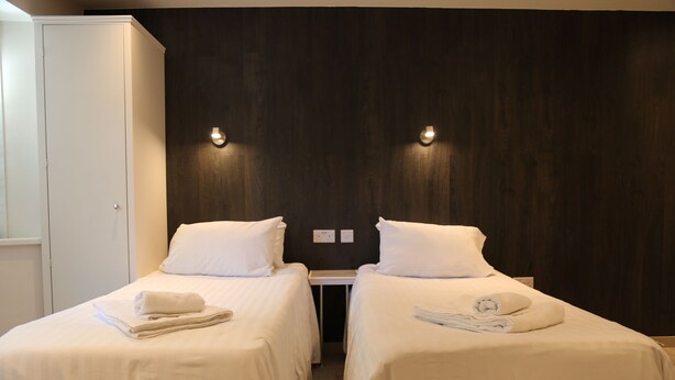 A hotel room with two single beds, a small table, and a wardrobe. - Debden Guest House (Uxbridge)