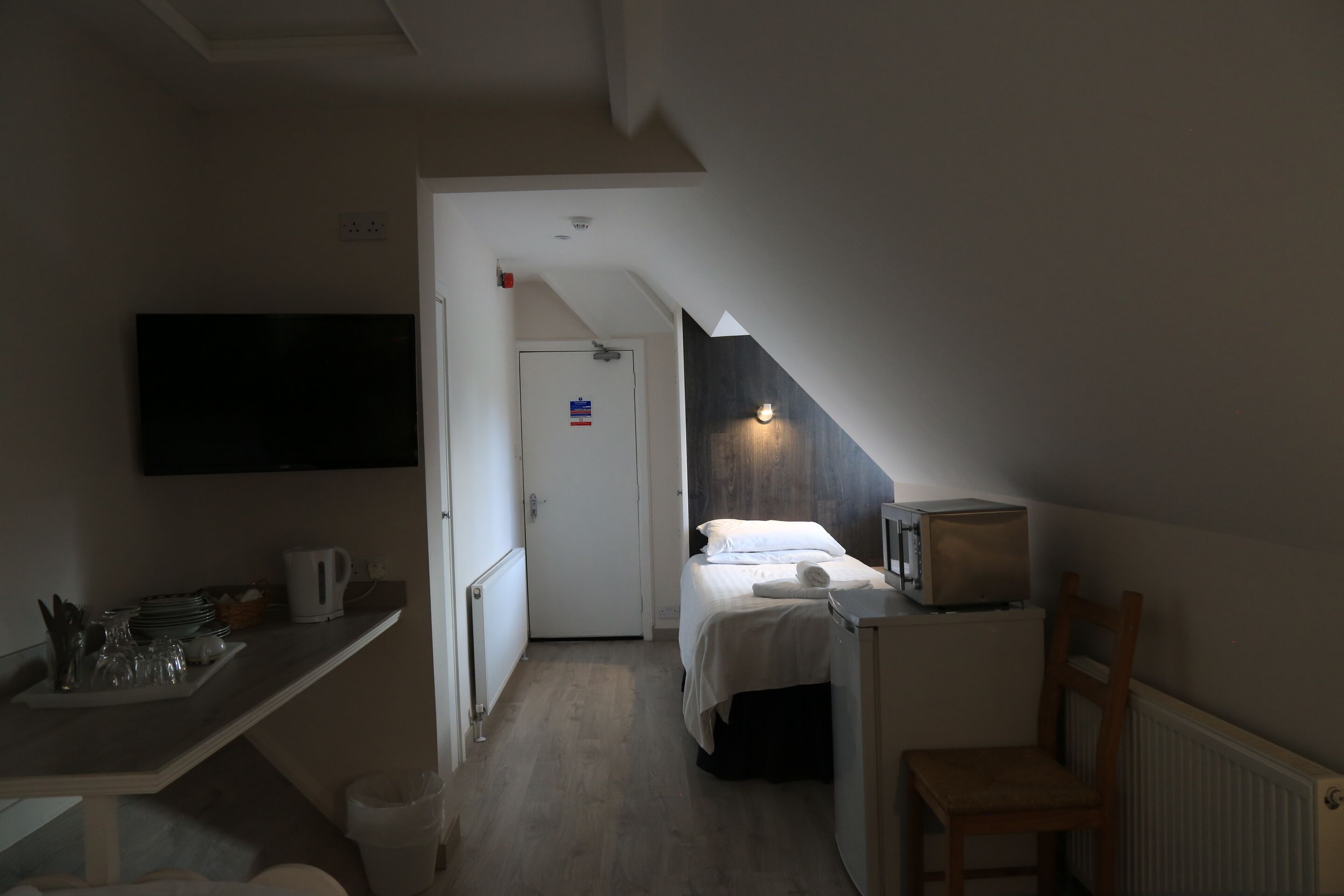 Une chambre d’hôtel de taille réduite, comprenant un lit, un petit bureau, une chaise, un four à micro-ondes et un téléviseur fixé au mur.
