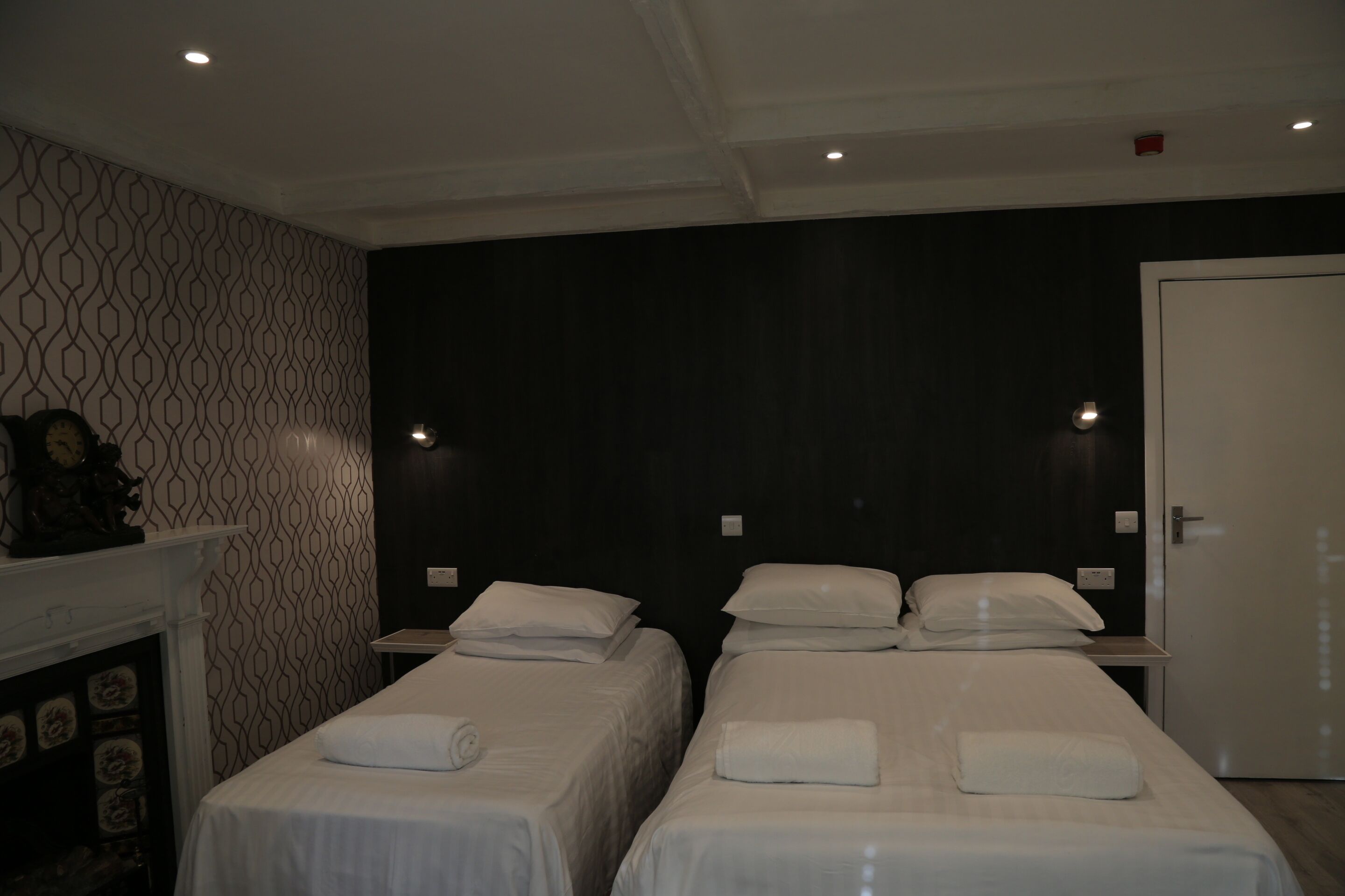 Quarto de hotel com duas camas de solteiro, lareira branca e parede escura com padrão geométrico.