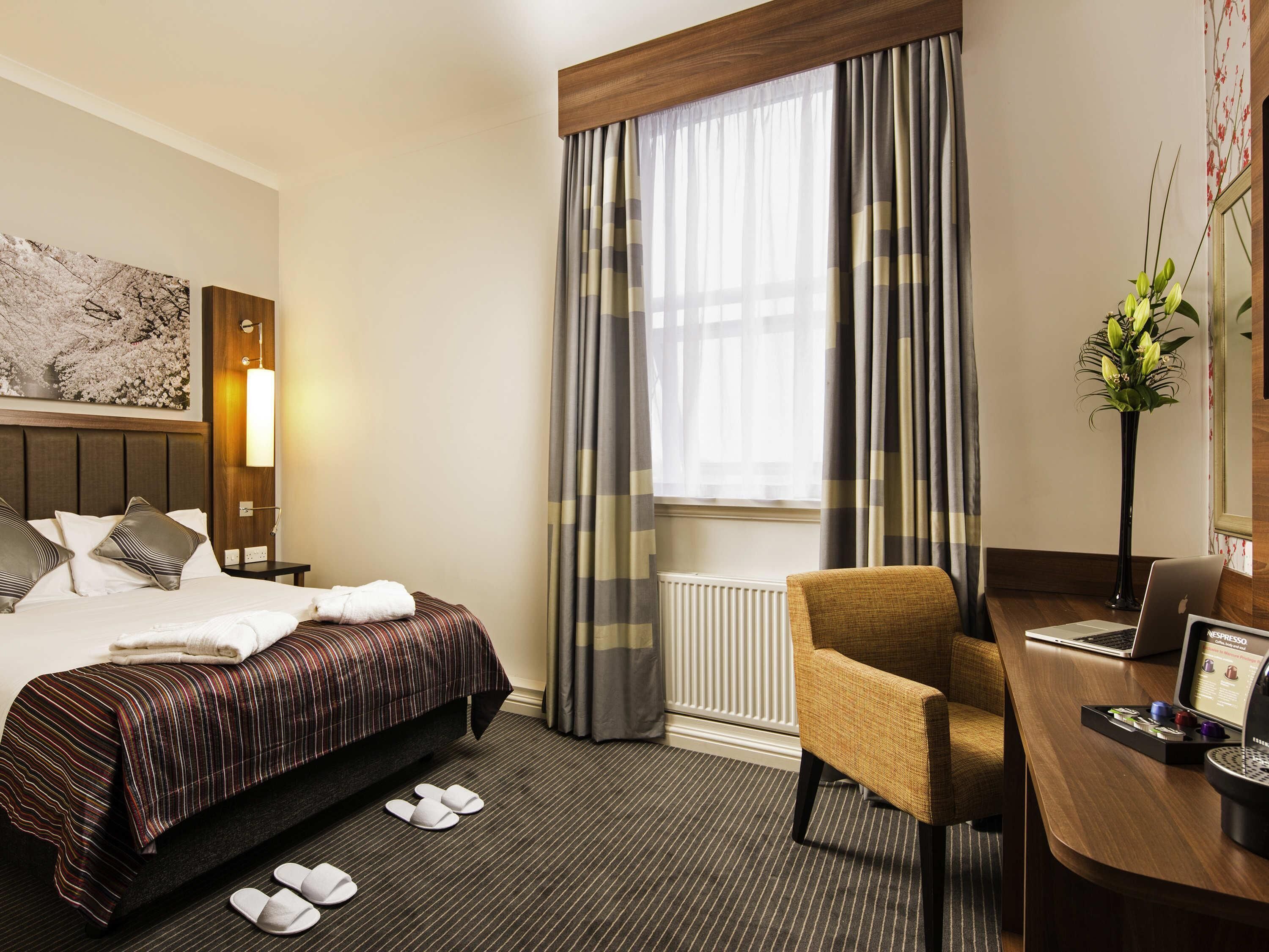 A hotel room with a bed, a desk with a laptop, a chair, a radiator, and a window with curtains.