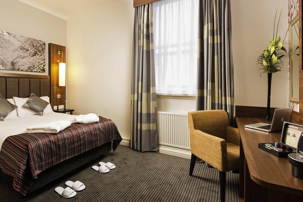 Une chambre d’hôtel avec un lit, un bureau avec un ordinateur portable, une chaise, un radiateur et une fenêtre avec des rideaux.