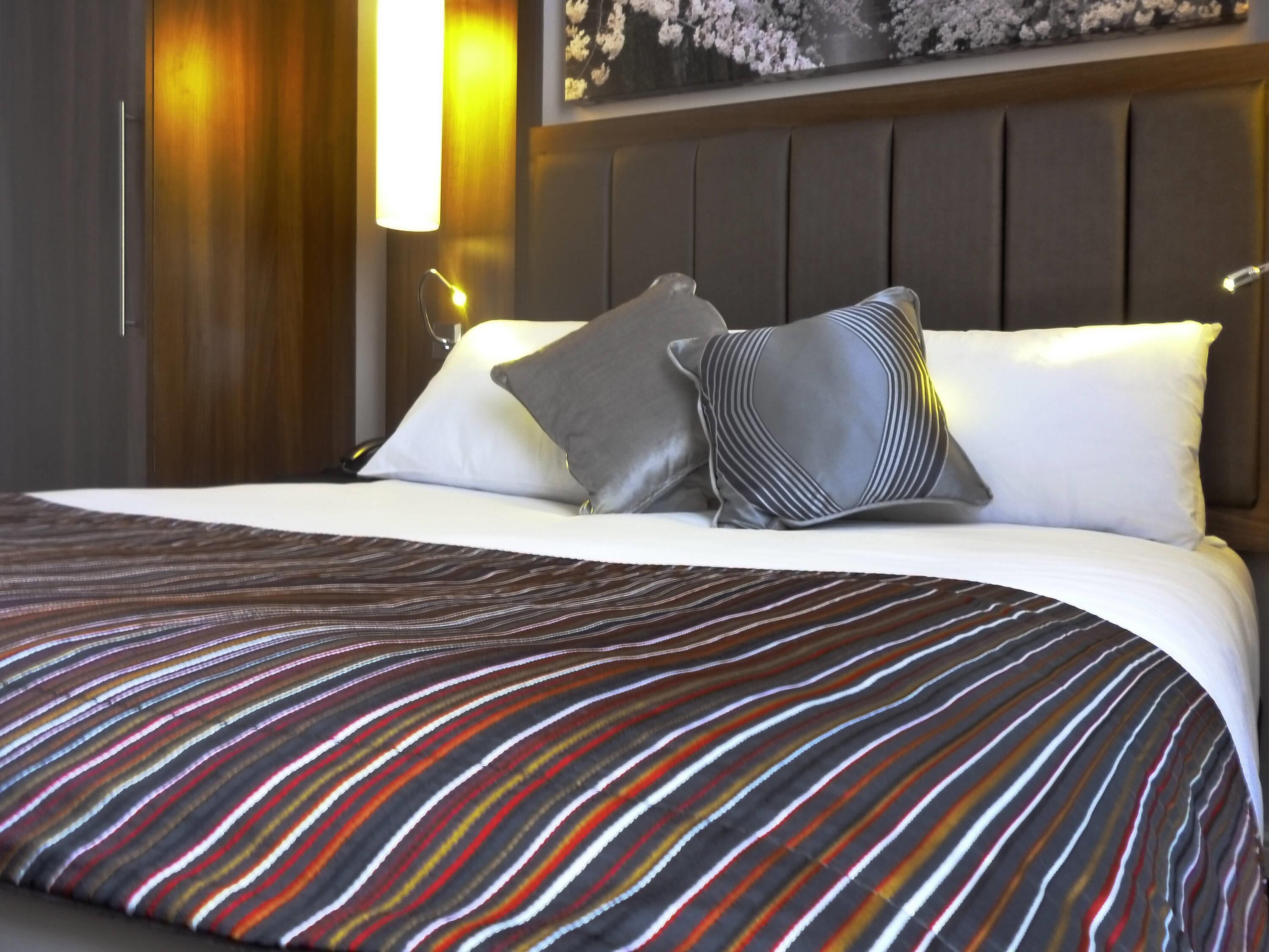 A hotel room with a bed, pillows, a striped bedspread, a wooden headboard, and a wall-mounted light fixture.