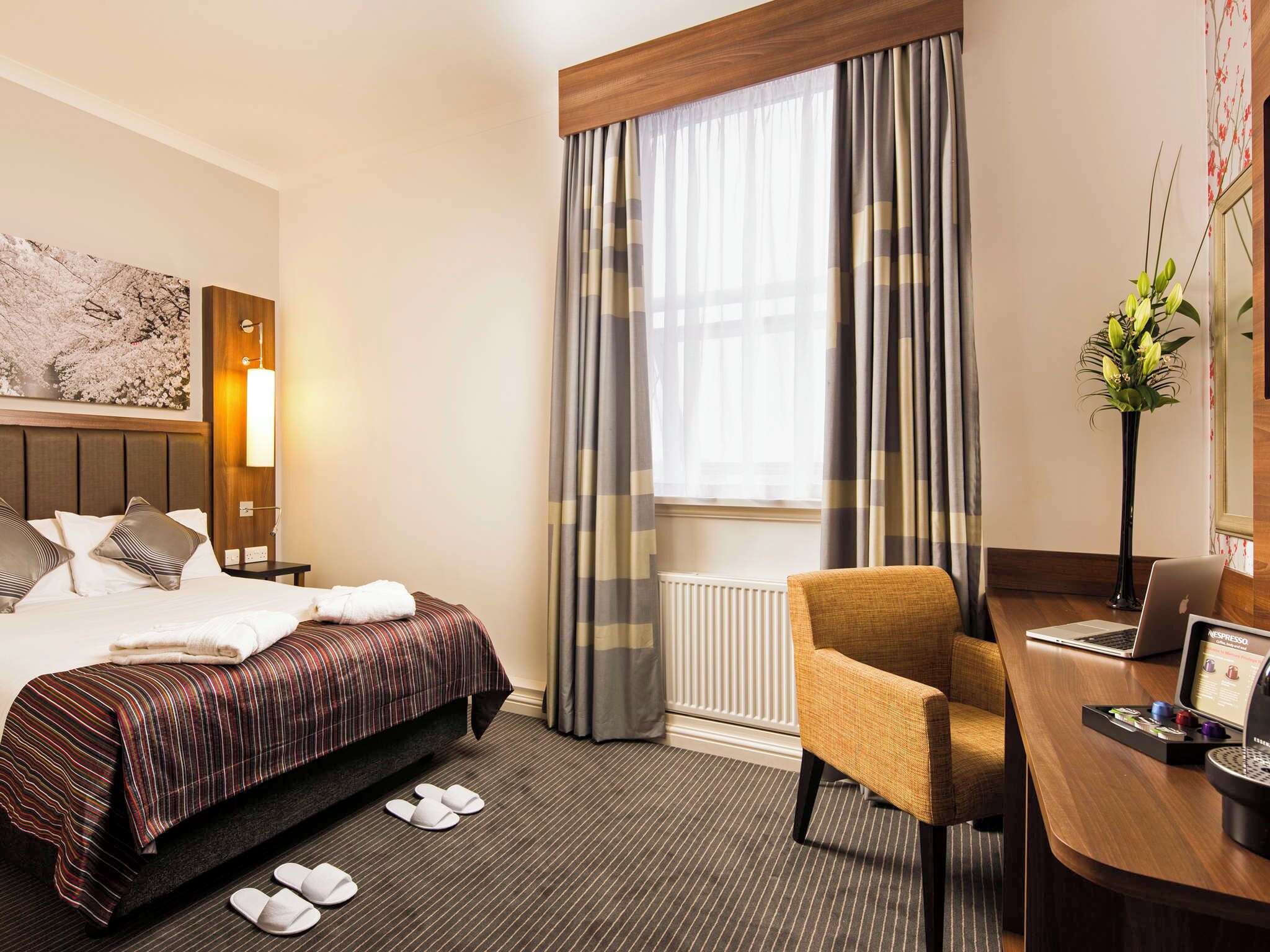 Une chambre d’hôtel avec un lit, un bureau avec un ordinateur portable, une chaise, un radiateur et une fenêtre avec des rideaux.
