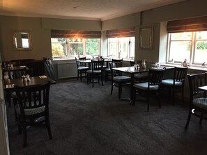 A dining area with multiple tables and chairs, large windows with curtains, and a radiator under the windows.