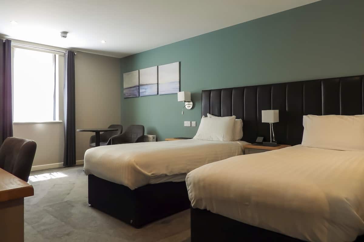 A hotel room with two beds, a desk, a chair, a radiator, and a window with curtains.