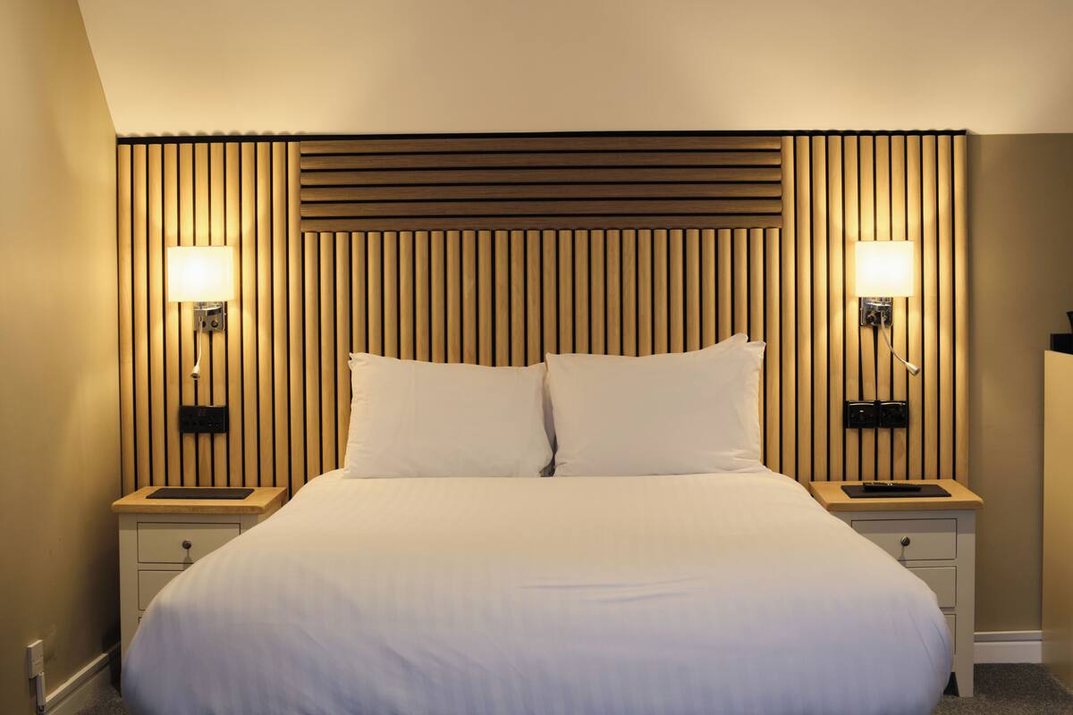 A hotel room with a large bed, two bedside tables with lamps, a wooden headboard, and a small drawer under each table.