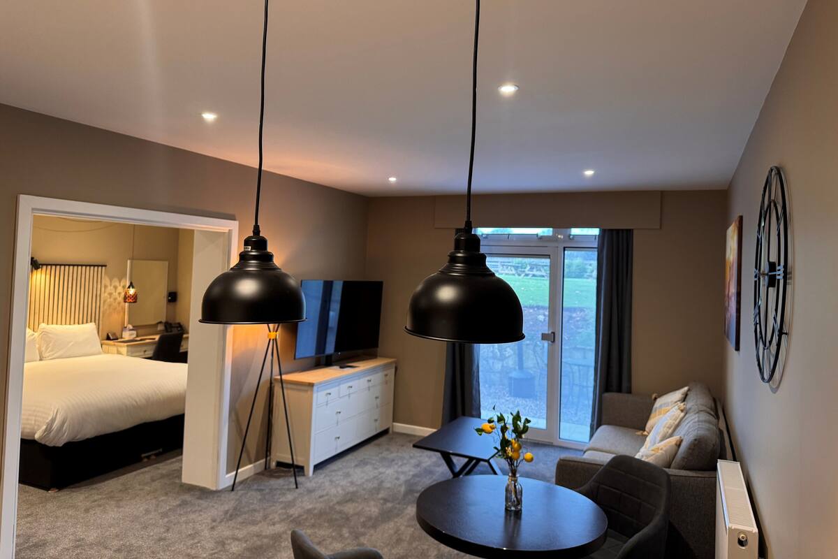 A modern hotel room with a bed, a desk, a TV, a sofa, and a dining table with a vase of flowers.
