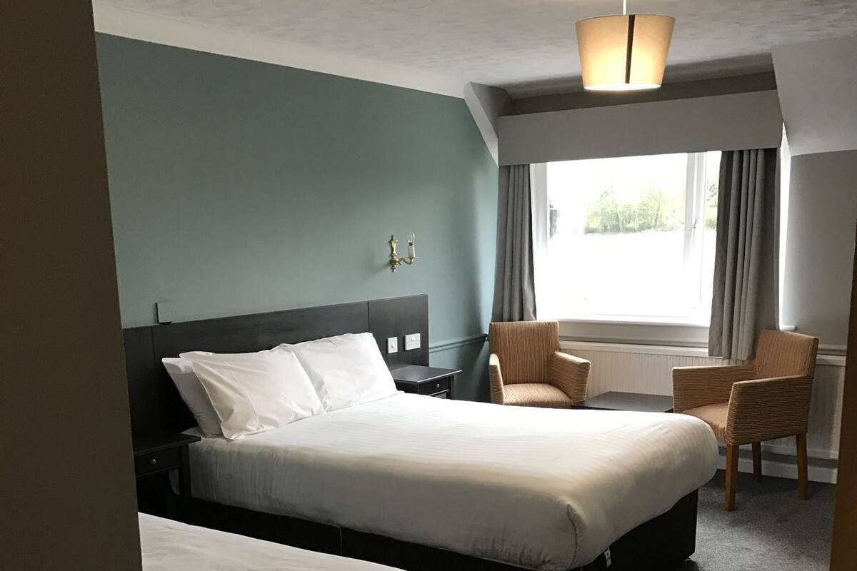 A hotel room with two beds, a chair, a lamp, and a window with curtains.