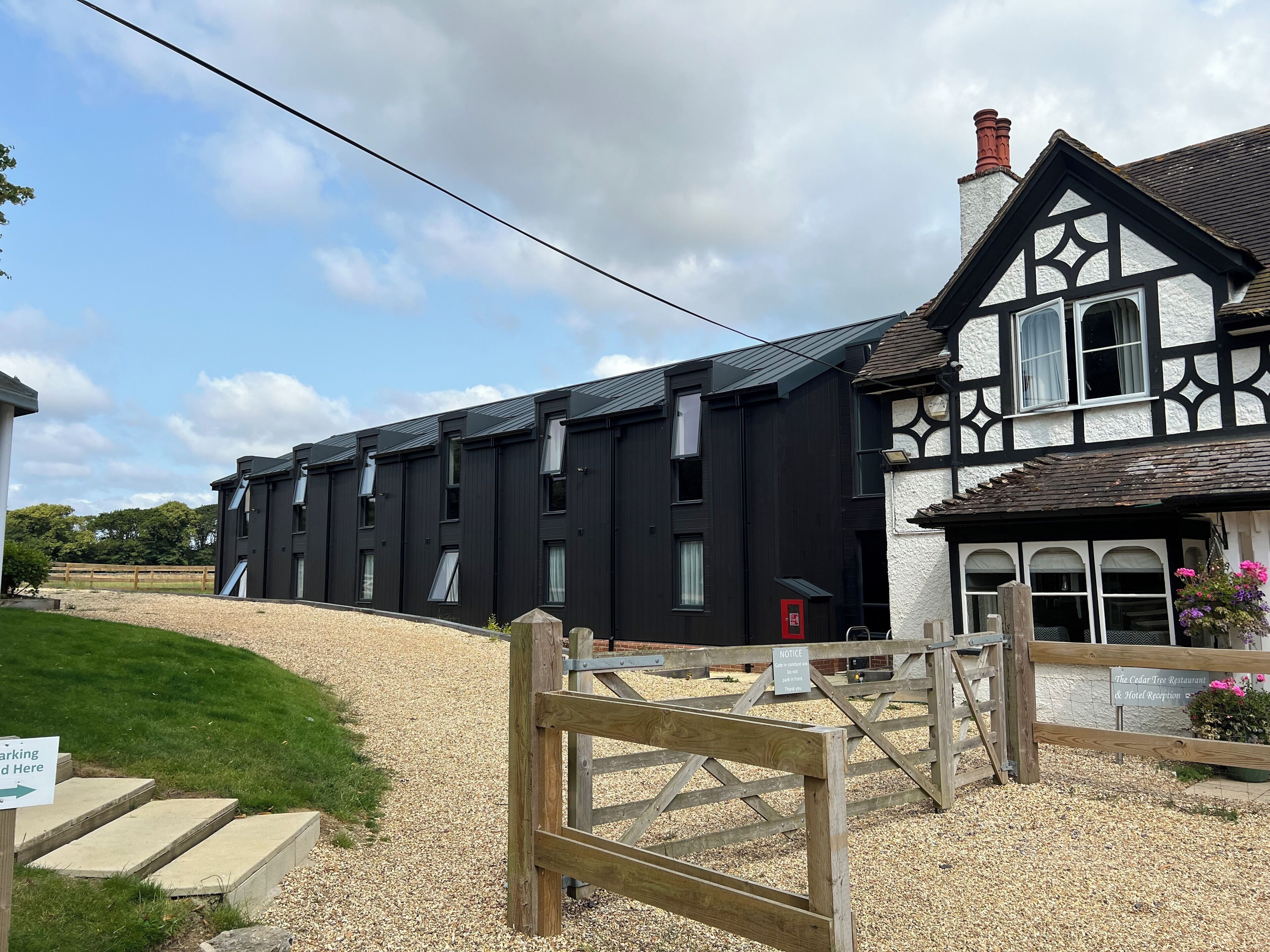 A modern building with a traditional half-timbered structure adjacent to it, a gravel driveway, and a wooden gate.
