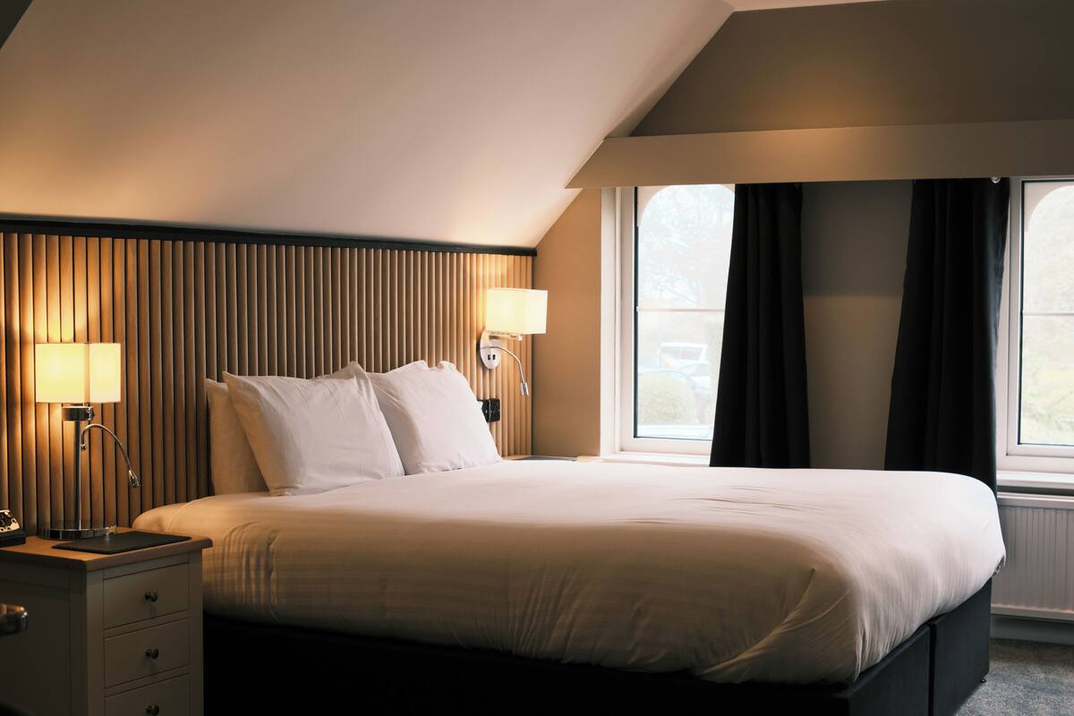 A hotel room with a bed, two bedside tables with lamps, a window with curtains, and a wall with vertical paneling.