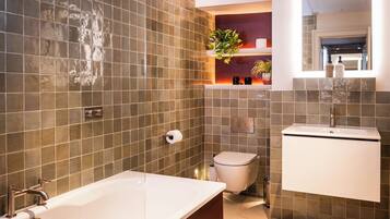 A modern bathroom with a bathtub, toilet, and sink, featuring a tiled wall and a shelf with plants.
