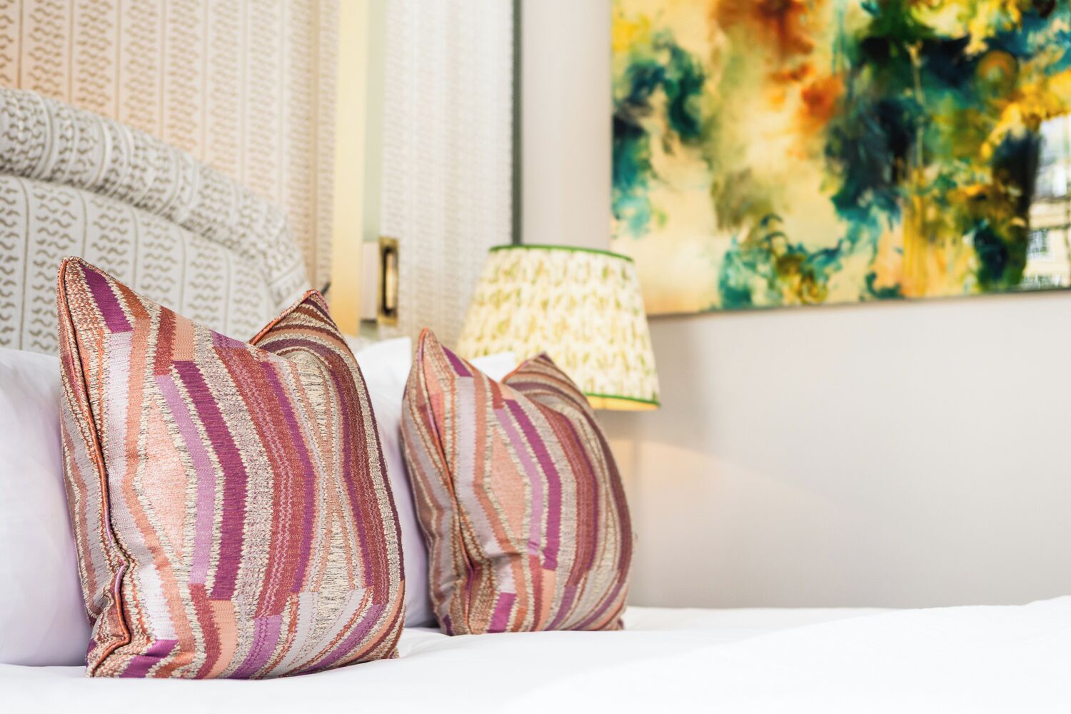 A neatly made bed with striped pillows and a patterned throw, a lamp, and a textured wall with a painting.