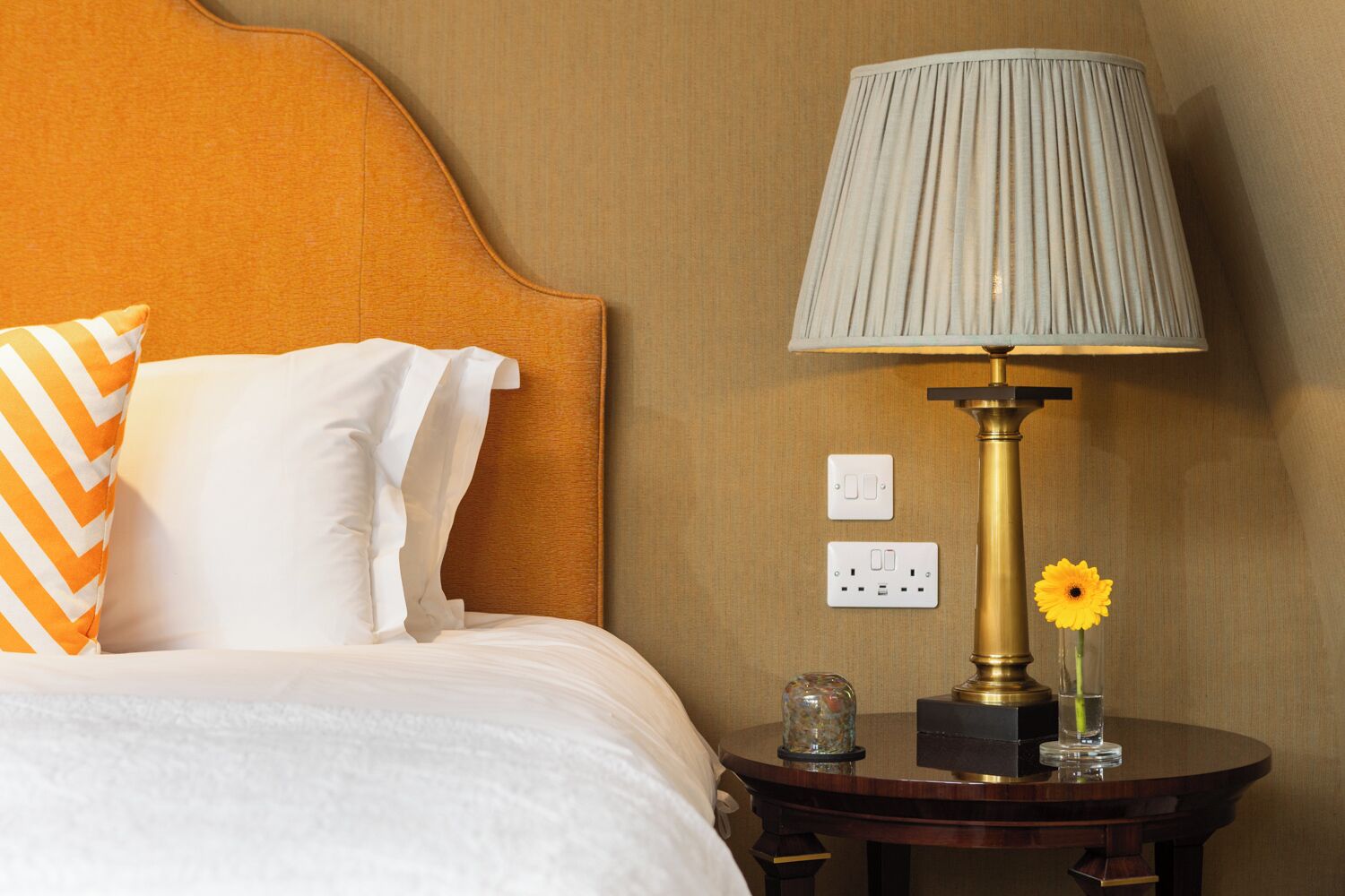 A hotel room with a bed, a nightstand with a lamp, a flower vase, and two electrical outlets.