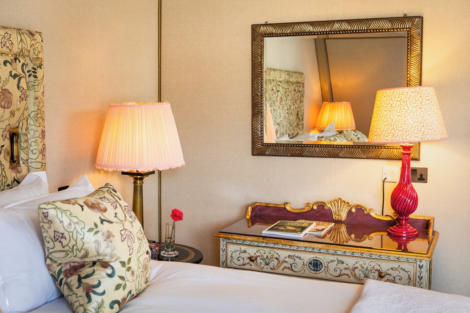 A hotel room with a bed, two bedside lamps, a mirror, a small table with a book, and a decorative chest.