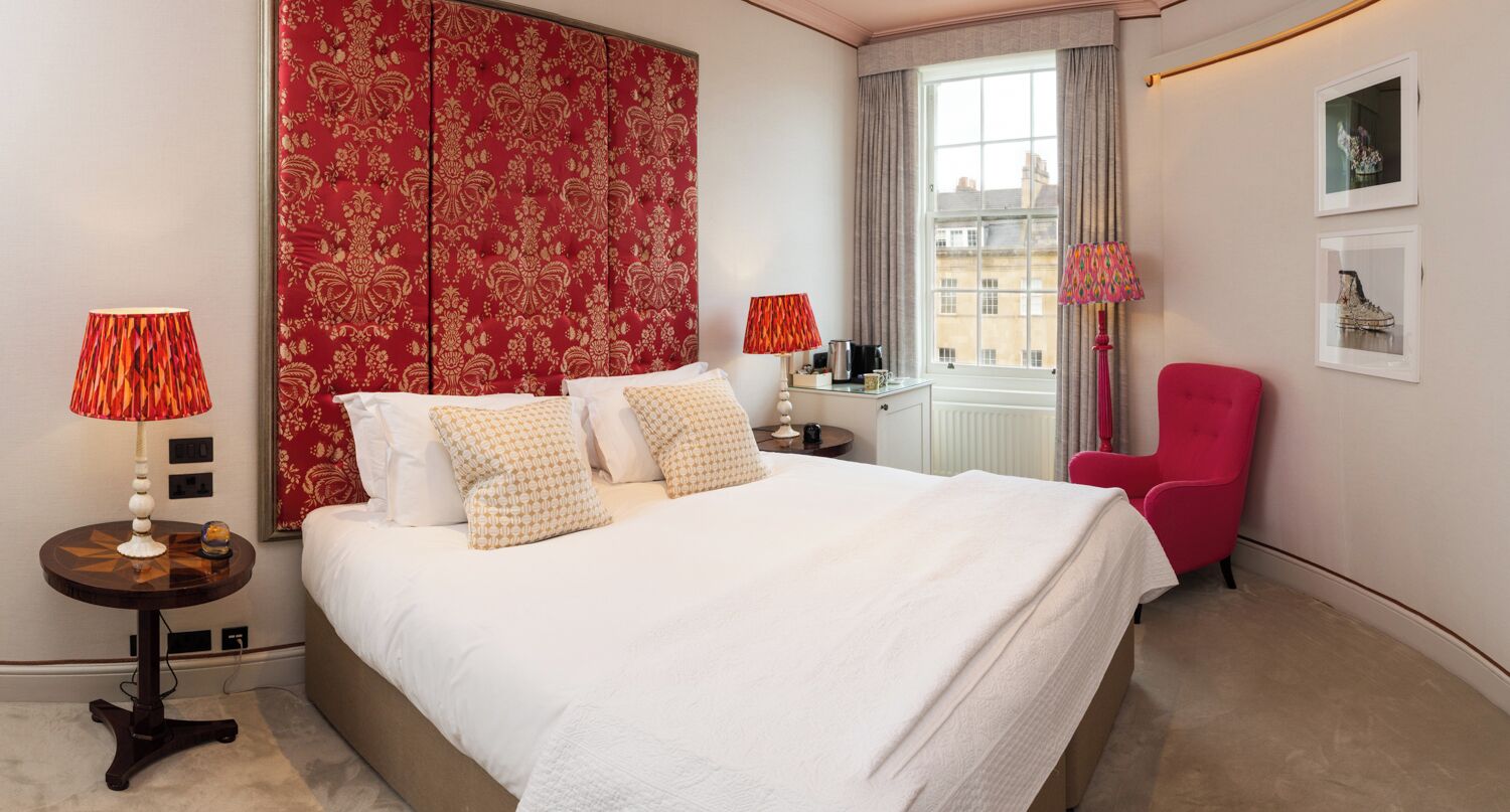 A hotel room with a large bed, two bedside tables, a red chair, and a window with curtains.