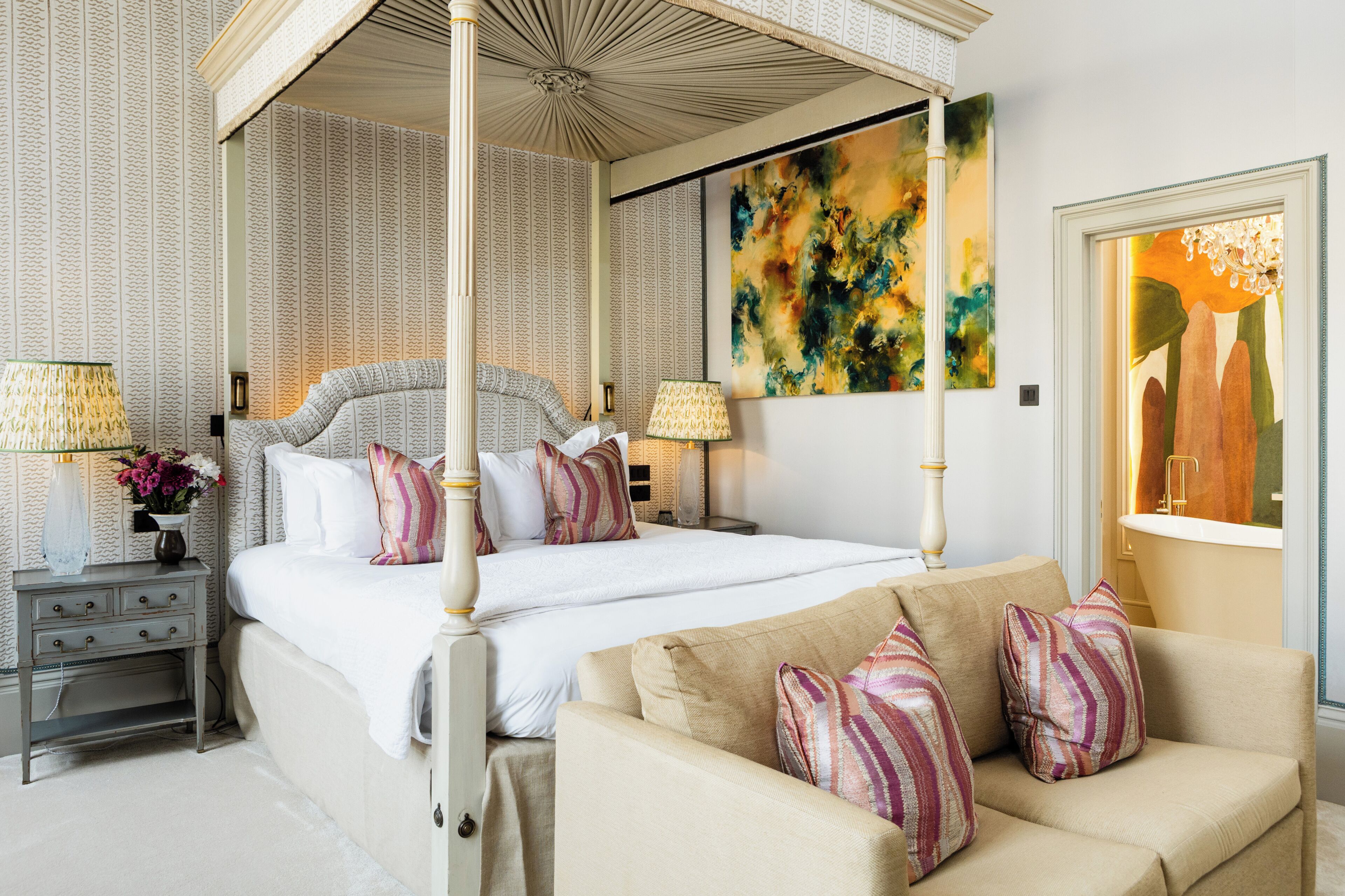 A four-poster canopy bed with multiple pillows, a bedside table with lamps, and a large abstract painting on the wall.