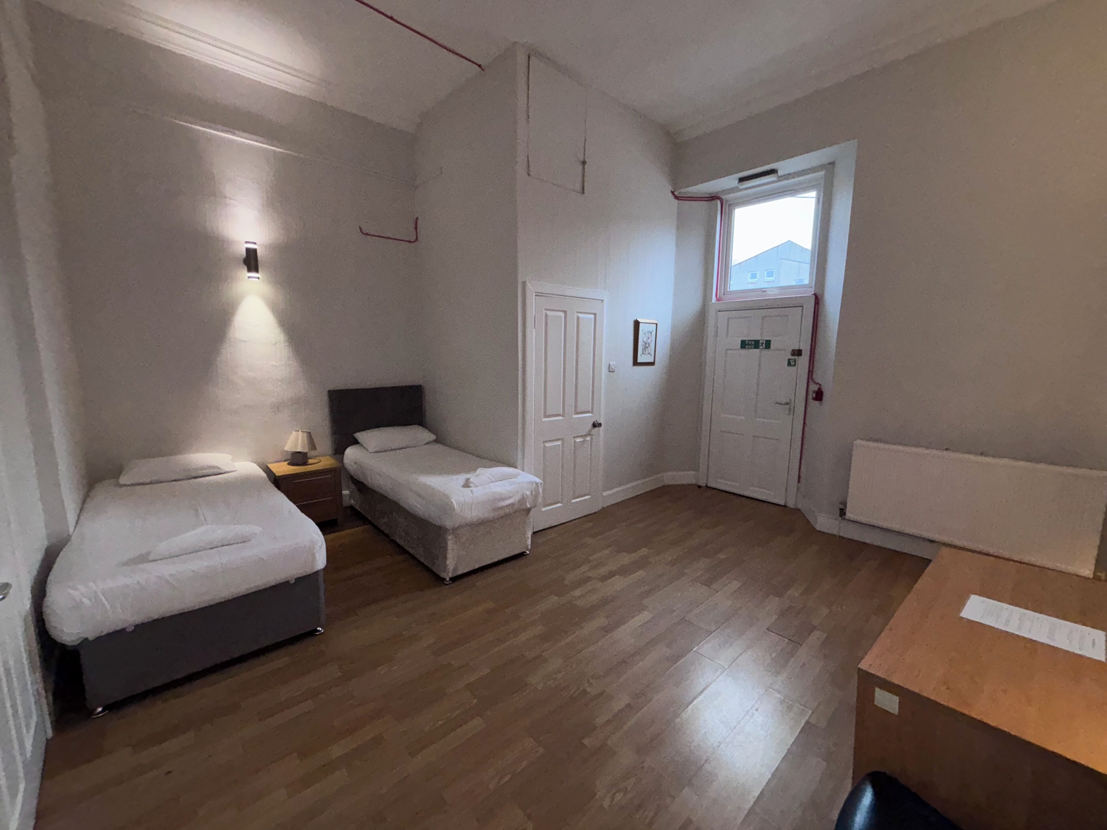 Una habitación pequeña y limpia con dos camas individuales, una mesita de noche, una lámpara de pared y una puerta que da a otra habitación.