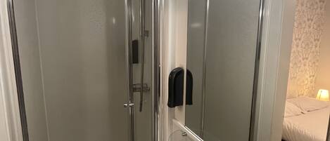 A modern bathroom with a glass-enclosed shower, a white sink, and a mirror reflecting a bed with a lamp.