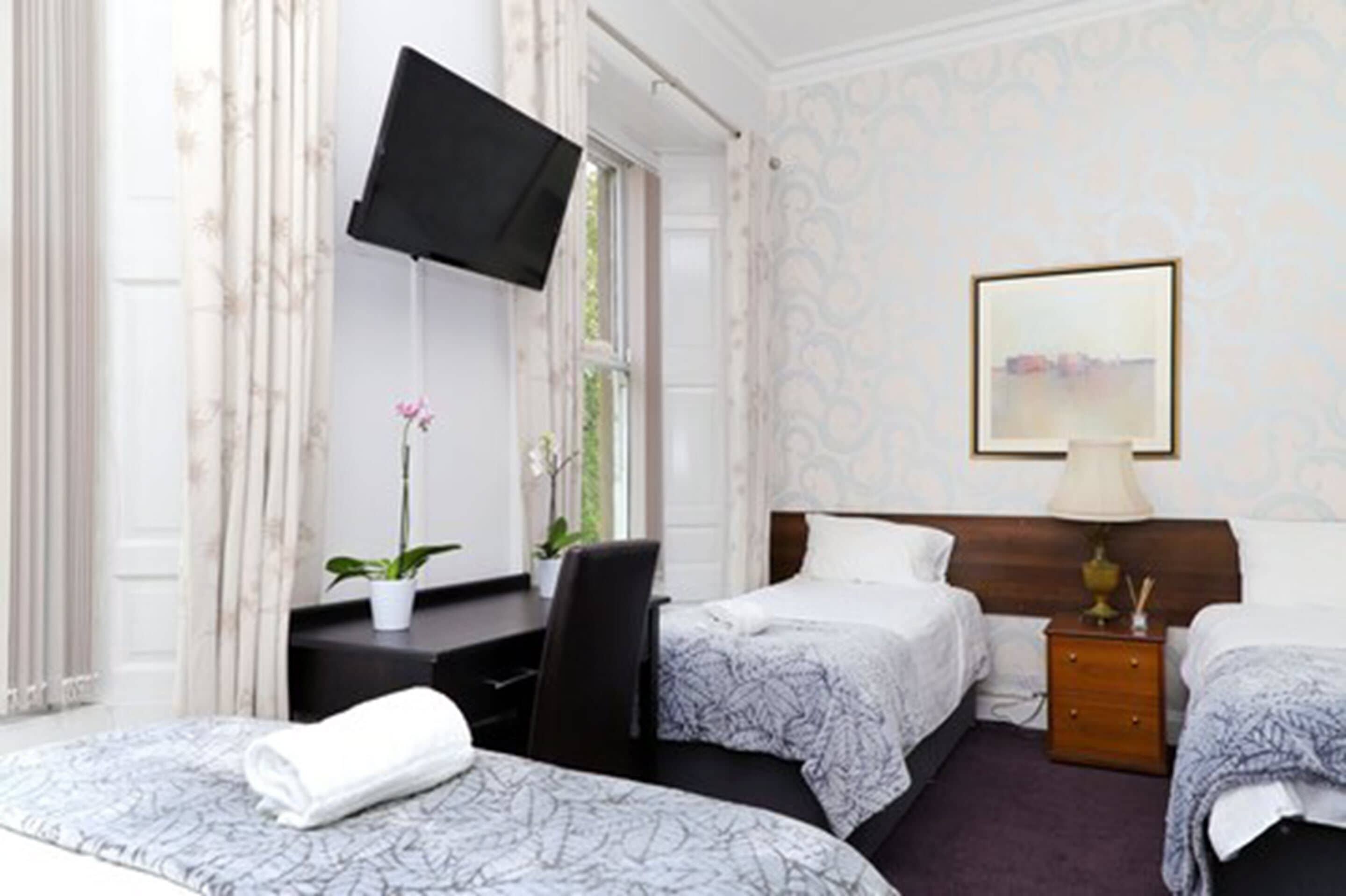 A hotel room with two beds, a desk, a TV, a bedside table, and a window with curtains.