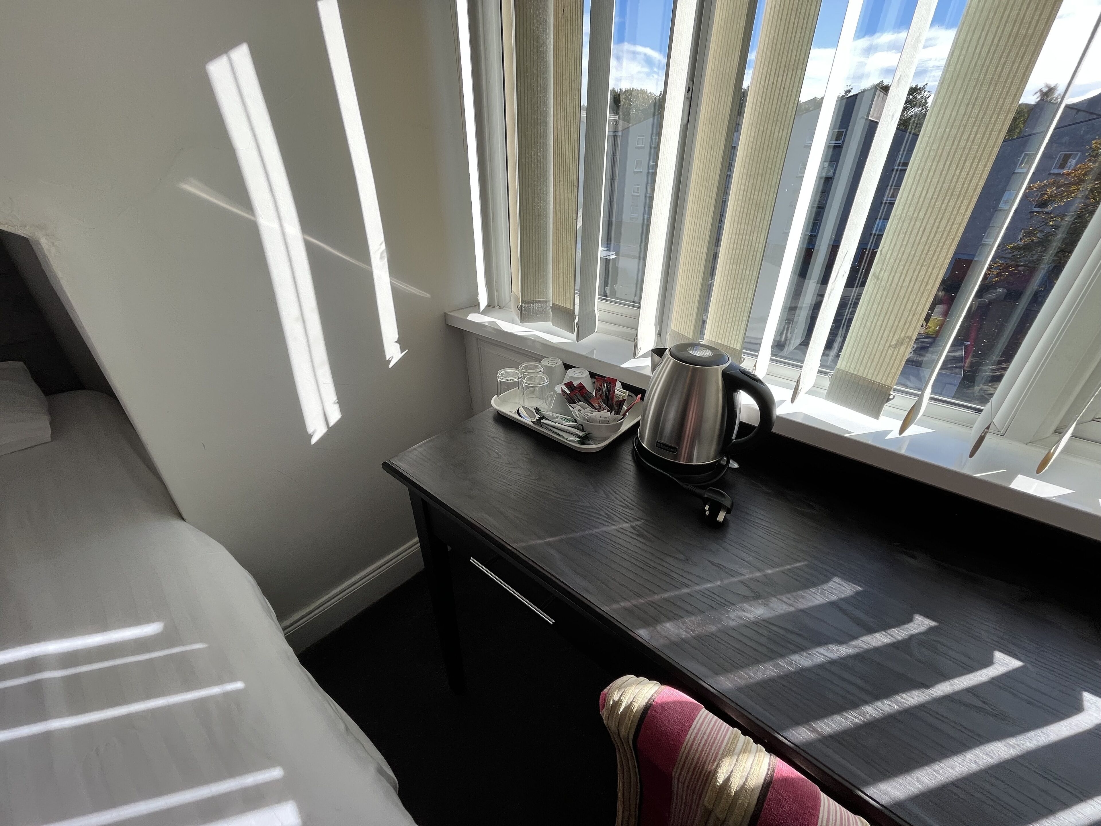A hotel room with a bed, a small desk with a kettle, a tray with cups, and a window with blinds.