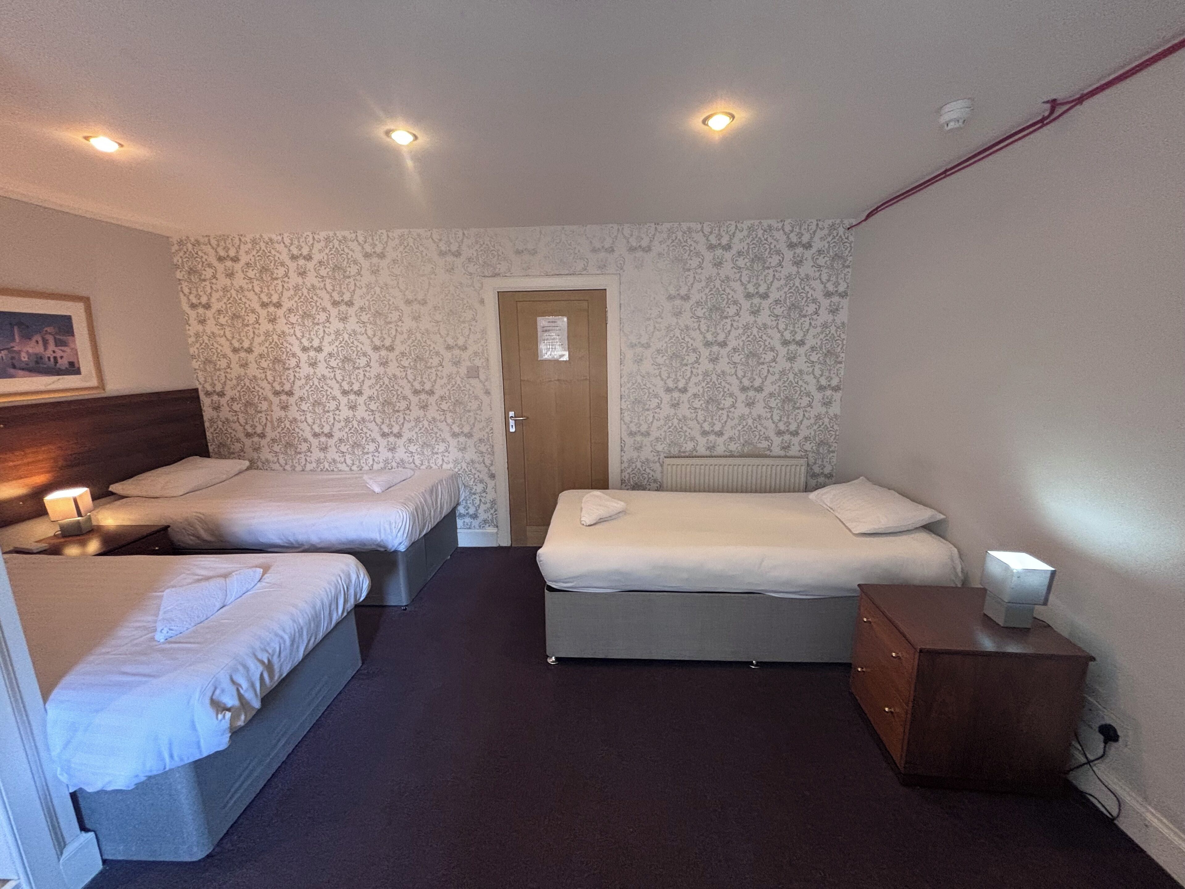 A hotel room with two single beds, a double bed, a wooden dresser, and a nightstand with a lamp.
