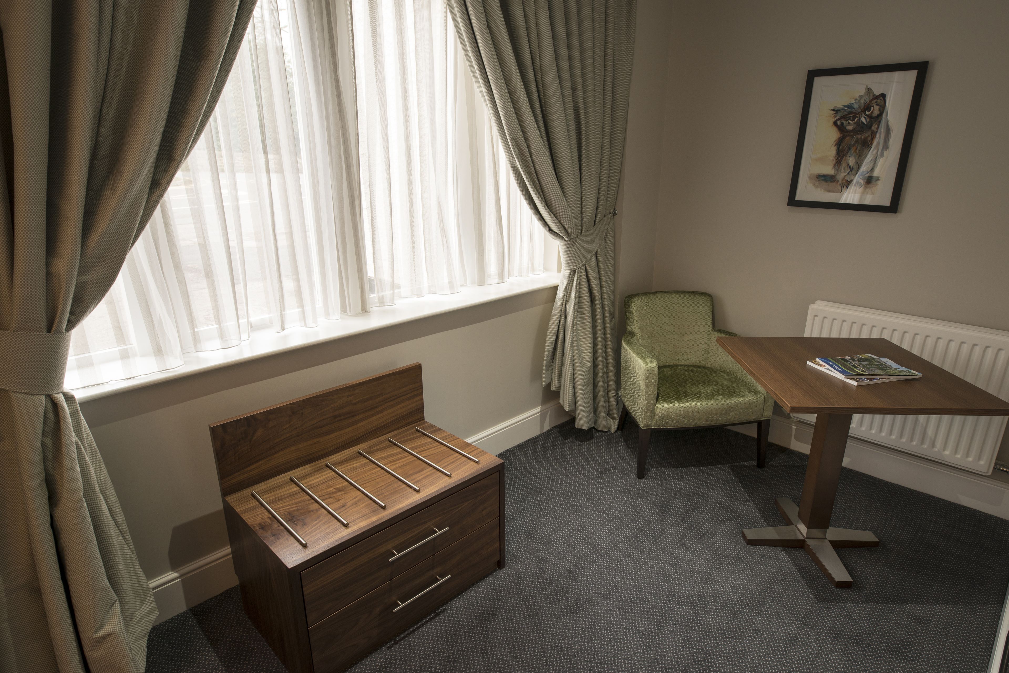 Un dormitorio con una cama de madera, una silla verde, una mesa con un libro y un ventanal con cortinas.