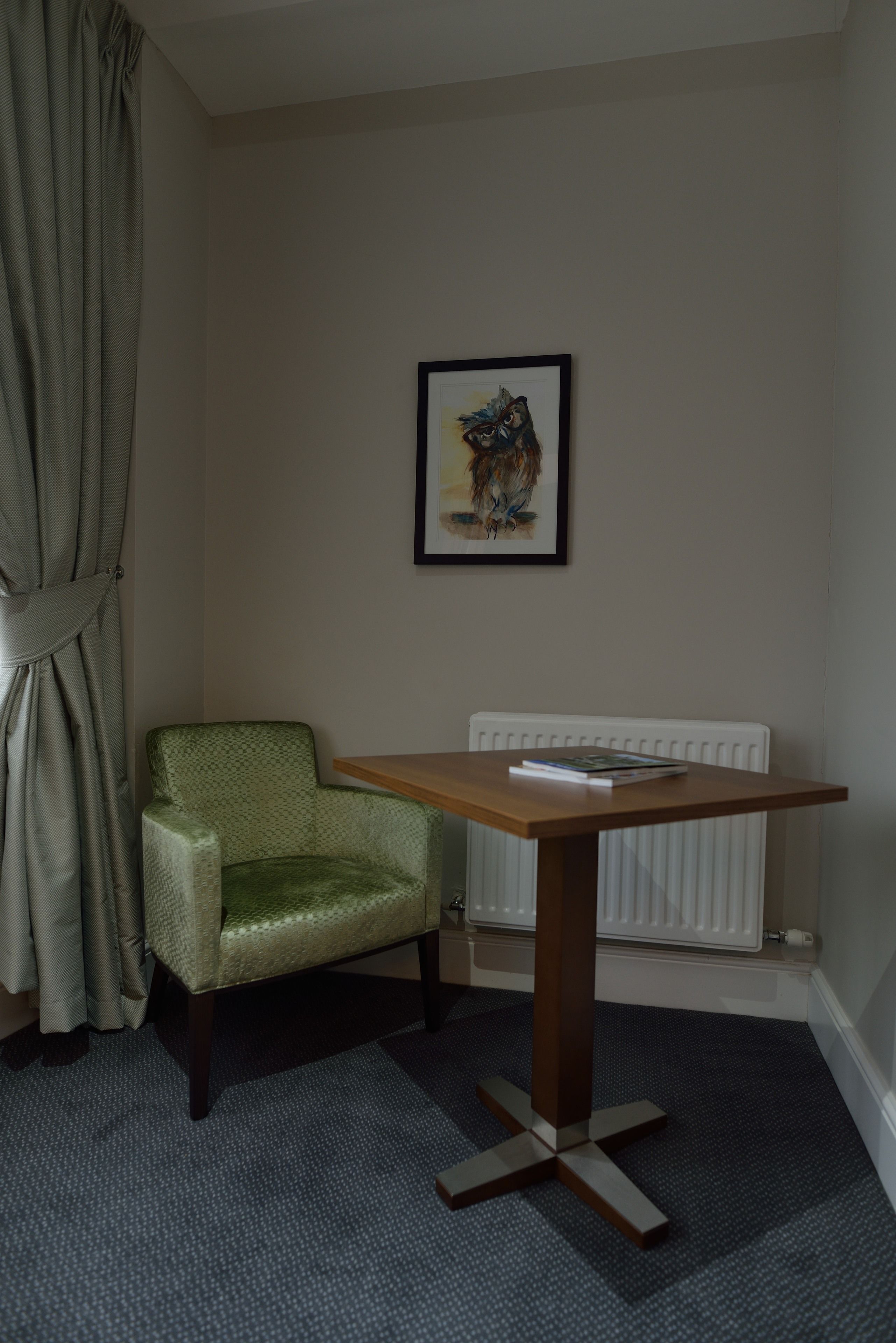 A room with a green armchair, a small wooden table, a framed artwork on the wall, and a radiator.