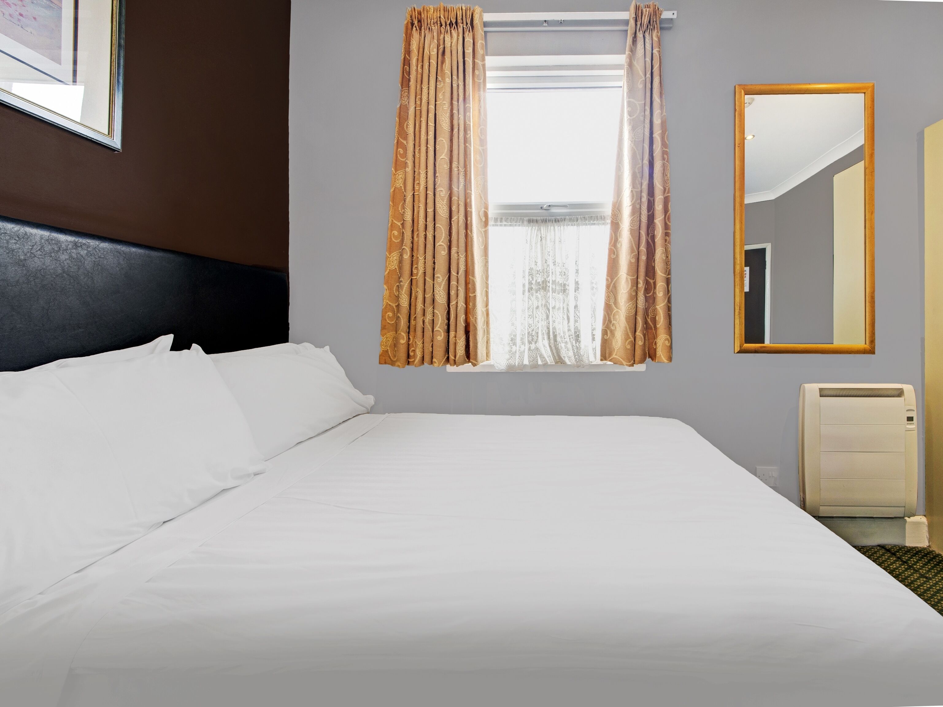 Een hotelkamer met een groot bed, een raam met gordijnen met een patroon, een spiegel en een radiator.