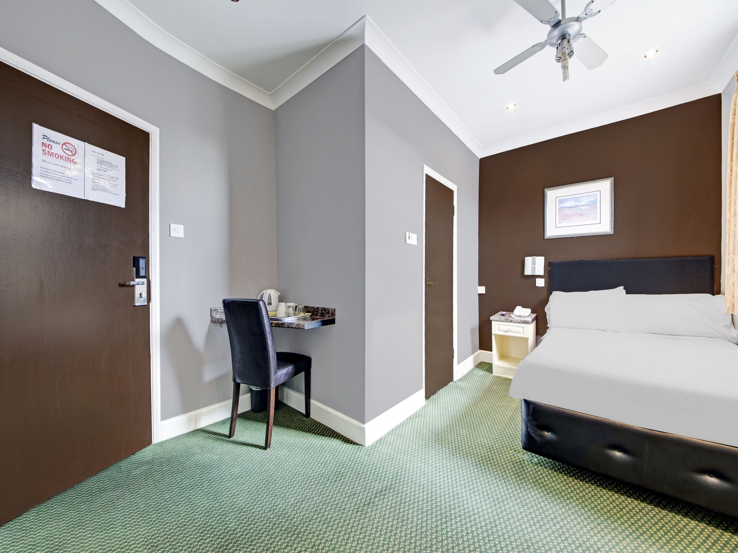 A hotel room with a bed, a desk with a chair, a ceiling fan, and a green carpet.