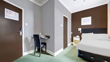 A hotel room with a bed, a desk with a chair, a ceiling fan, and a green carpet.