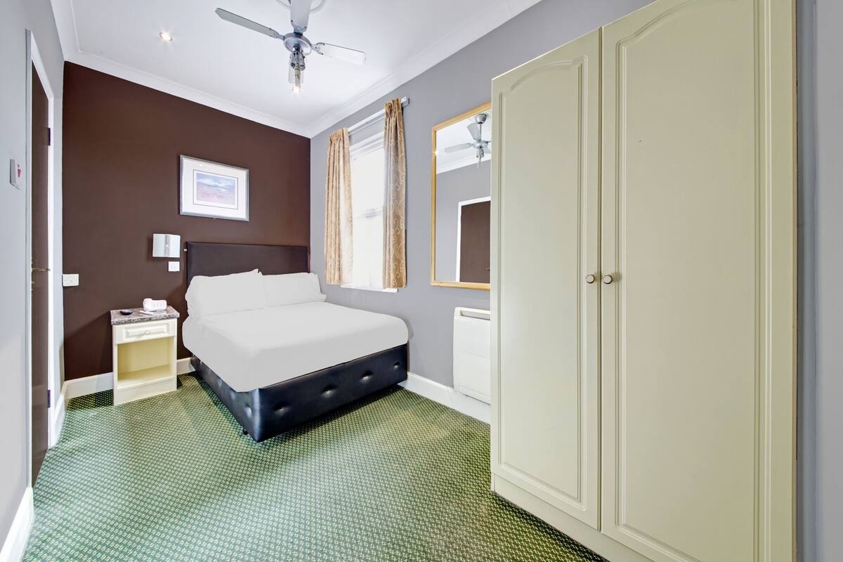 A hotel room with a bed, a ceiling fan, a nightstand, a mirror, and a wardrobe.