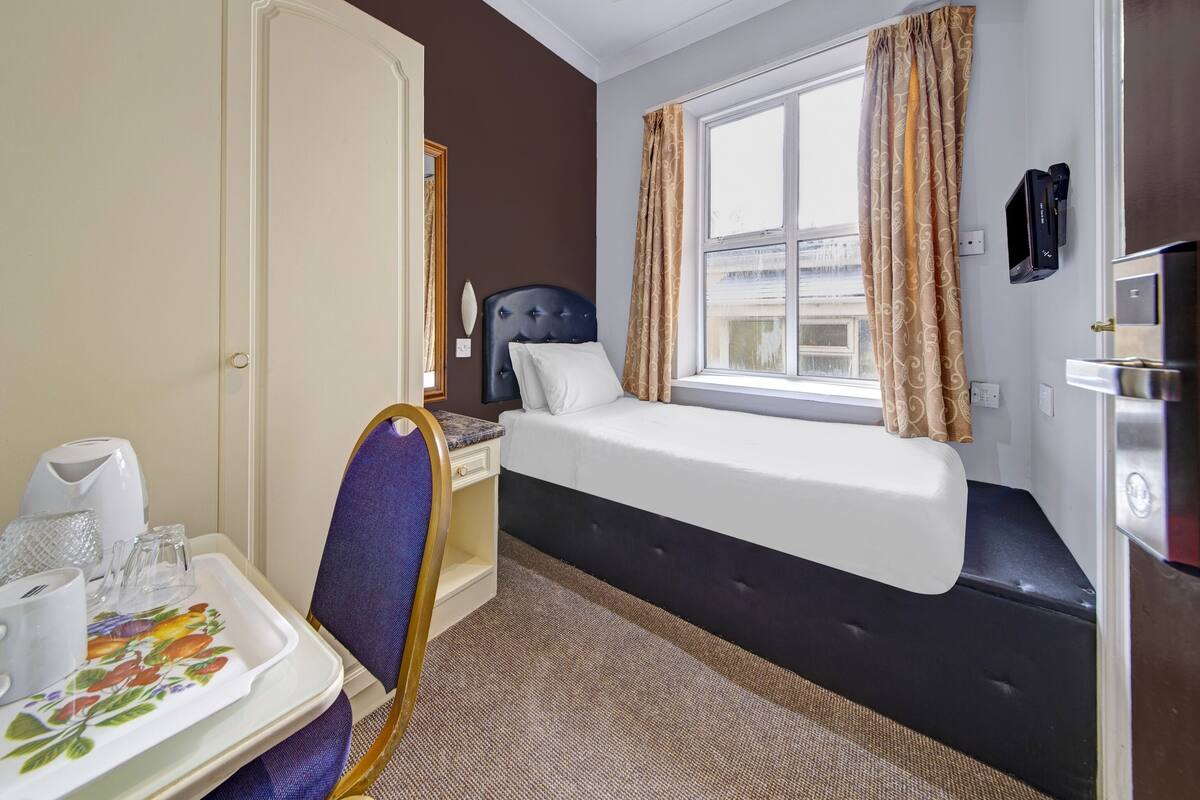 A compact hotel room with a bed, a desk with a chair, a small table with a tray, and a window with curtains.