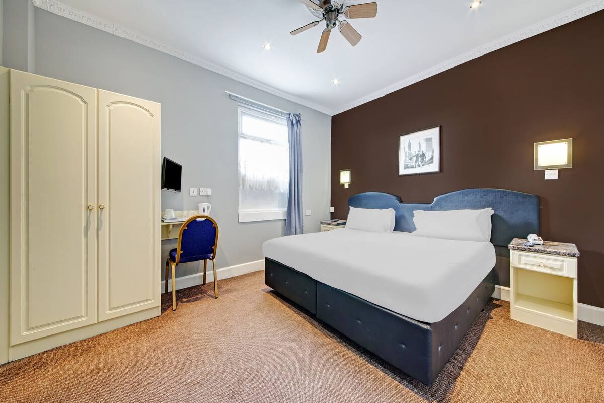 A hotel room with a large bed, a desk, a chair, a wardrobe, a ceiling fan, and a window with curtains.