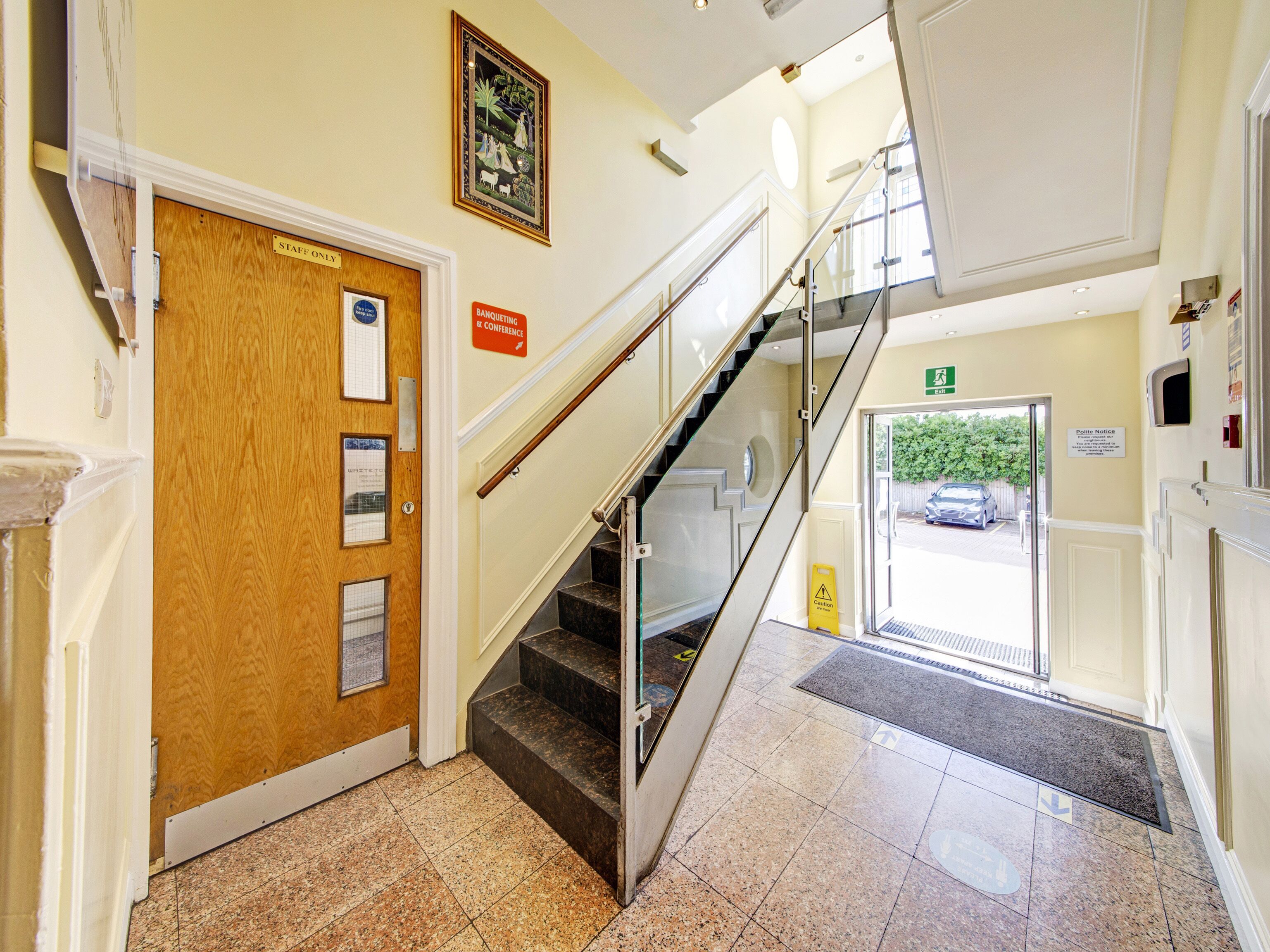Una hall d'albergo con una porta di legno con la scritta “STAFF ONLY”, una scala con ringhiera in vetro e un cartello con la dicitura “POLITE NOTICE”.