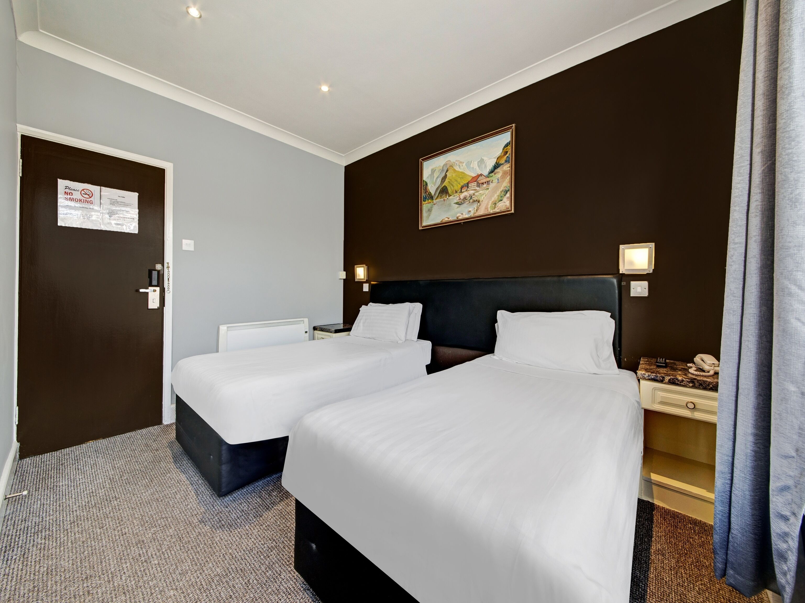 Habitación de hotel con dos camas, un cuadro en la pared, una puerta con letrero de no fumar y una mesita de noche con un teléfono y una lámpara.