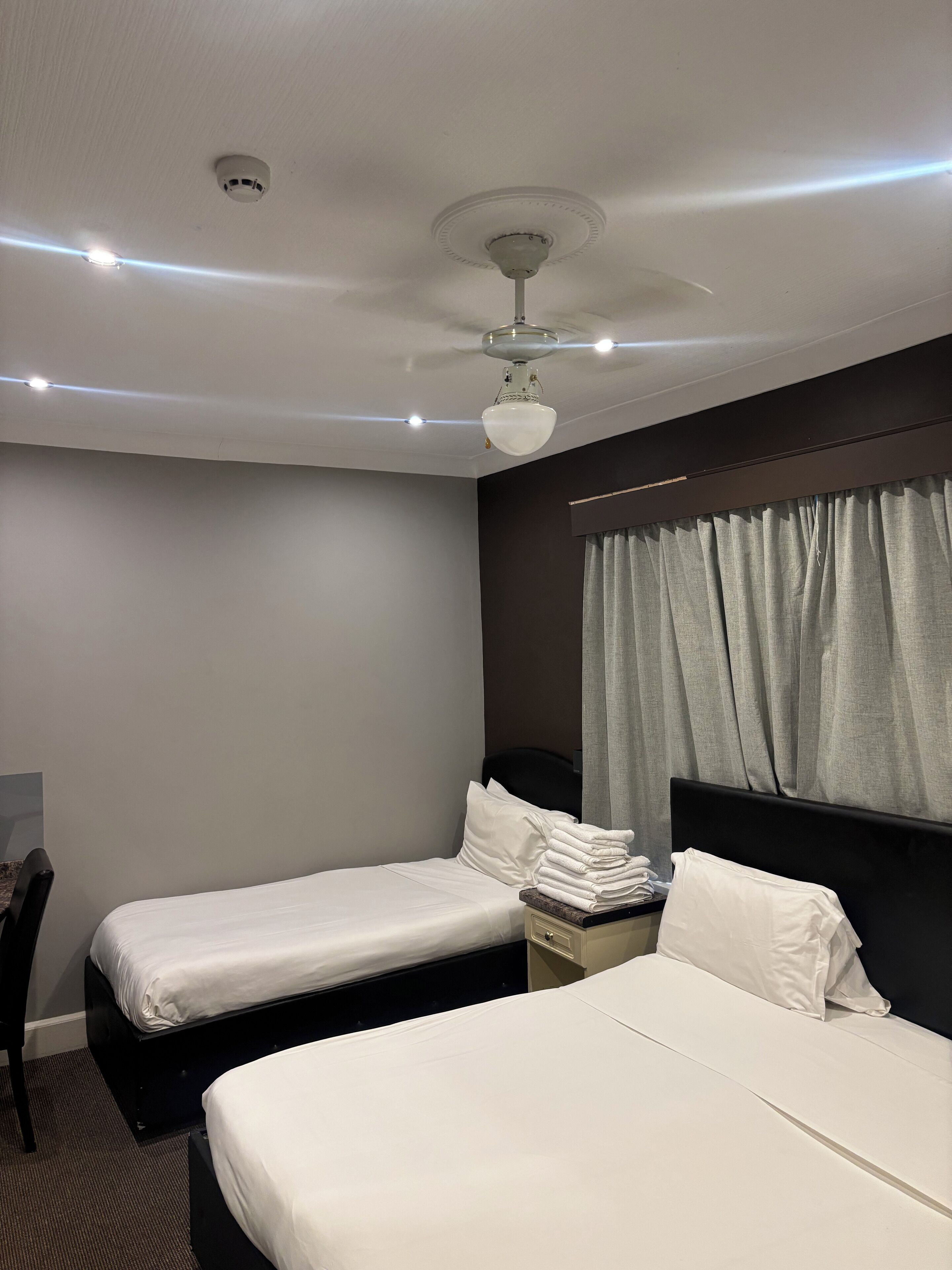 Ein Hotelzimmer mit zwei Einzelbetten, einem Deckenventilator, einem Nachttisch, einem Sessel und einem fensterbepflanzten Fenster.